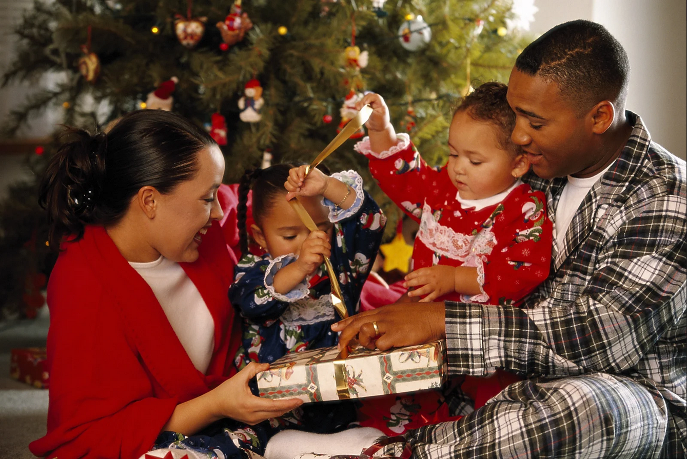 Parents helping two children open presents in front of the Christmas tree.